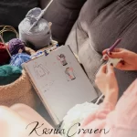 Designer's hands crocheting next to a notebook with sweater sketches and colorful yarn in a cozy freelance artist studio.